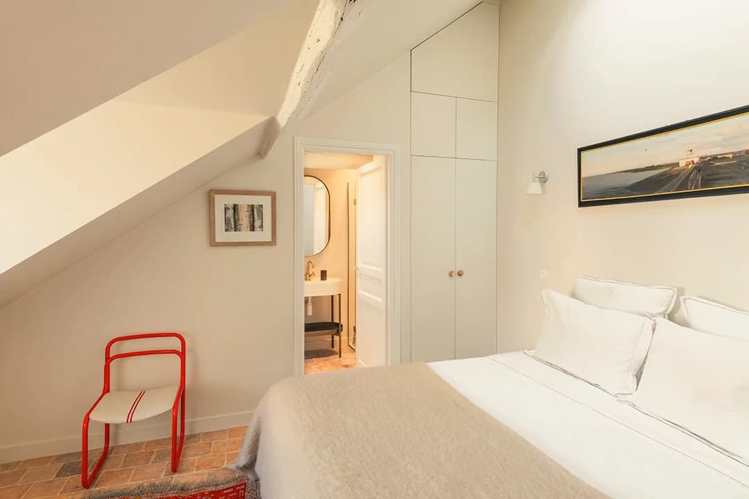 Eaved bedroom with red metal chair, ensuite glimpse and seascape art