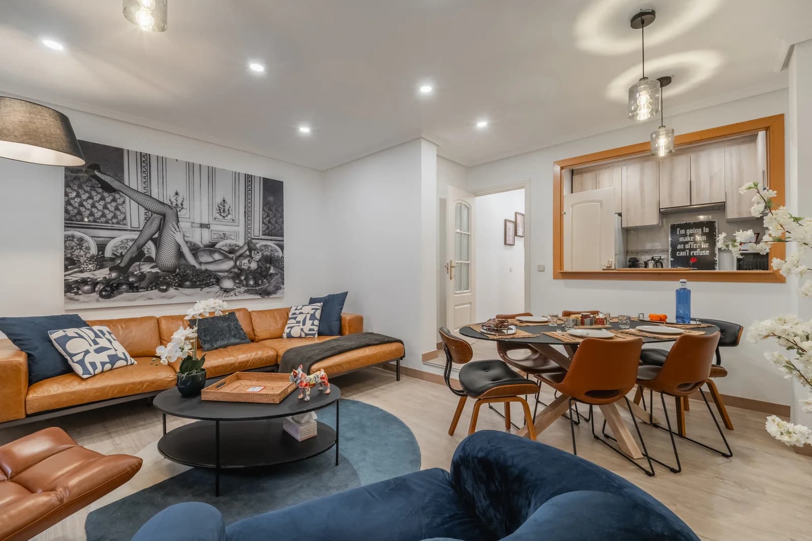 Open-plan living and dining with tan leather sofa, blue rug, kitchen pass-through