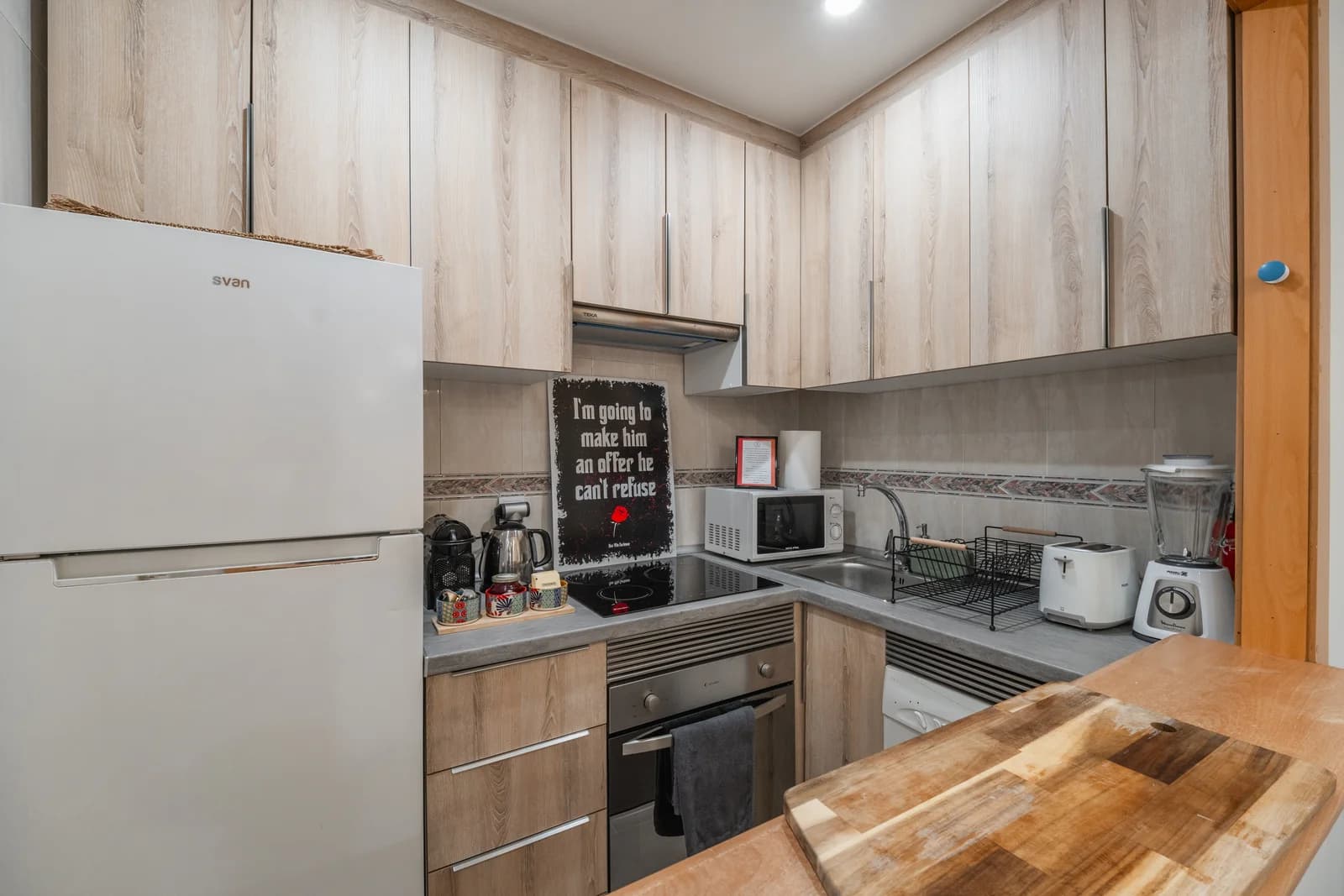 Compact light-wood kitchen with full-size fridge and stone countertops