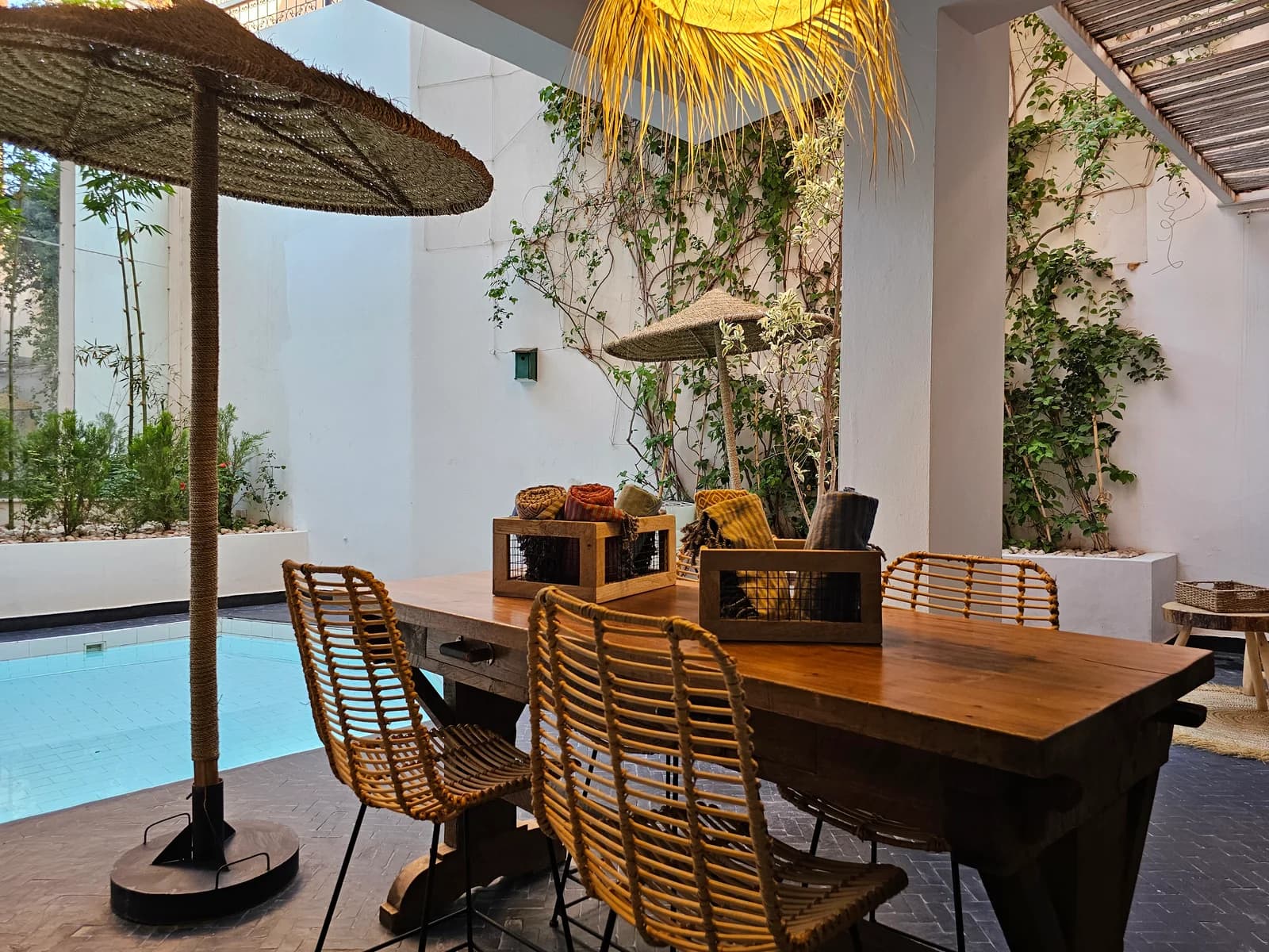 Outdoor dining table beside pool under thatched parasol and climbing greenery
