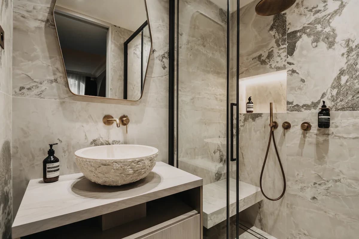 Stone basin on marble vanity beside walk-in shower with brass fittings