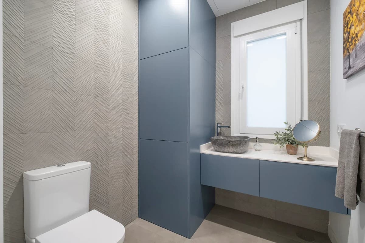 Slate-blue vanity beneath window with stone basin and herringbone tile